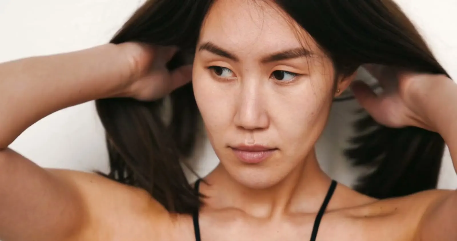 흰 벽 앞에서 두 손으로 긴 흑발을 쓸어 넘기는 모델